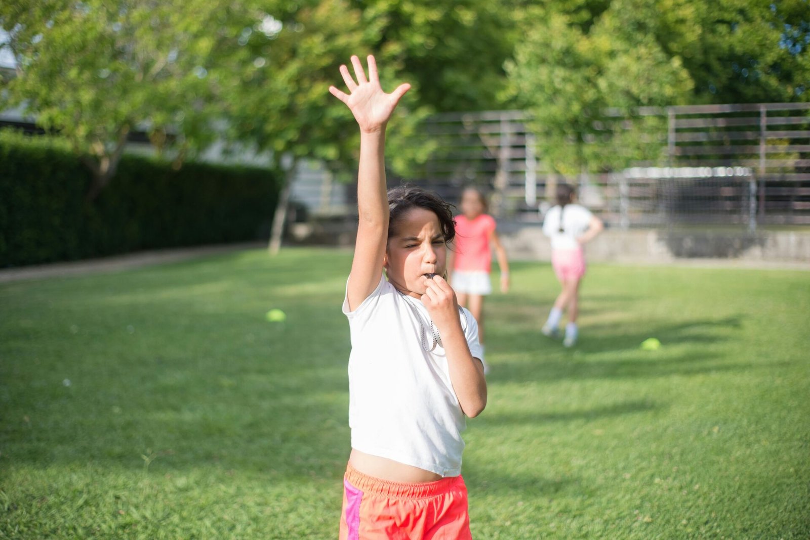 A young girl raises her hand and blows a whistle in a sunny outdoor setting with other children in the background.